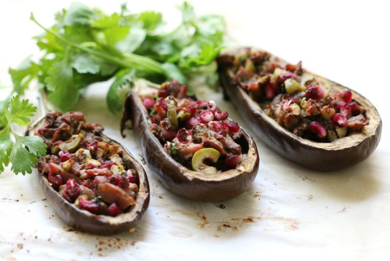 Three halved and roasted eggplants are filled with a colorful mixture of diced vegetables and pomegranate seeds. Fresh cilantro is placed behind them on a light-colored surface.